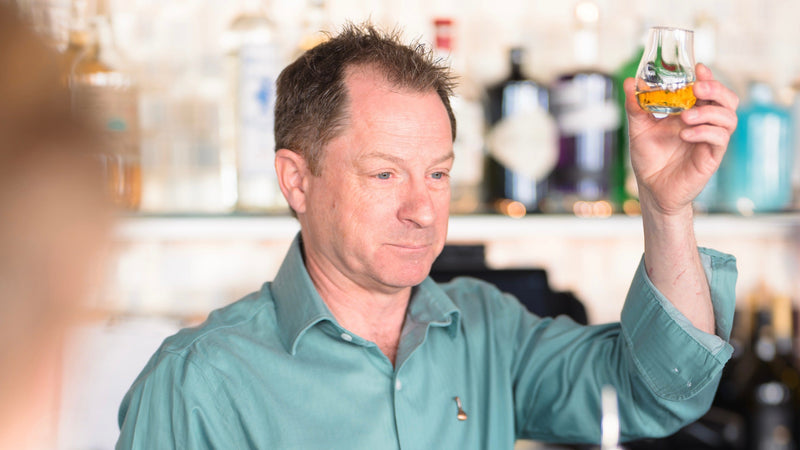 Man in a bar wearing a green shirt holding a small glass in the air in his left hand.