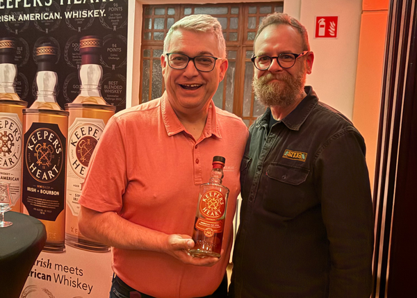Two men standing next to each other holding a bottle of Keeser's Heart whiskey in front of a promotional poster.