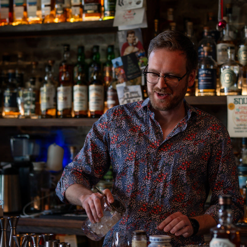 Man behind a bar preparing a drink with bottles and bar equipment in the background
