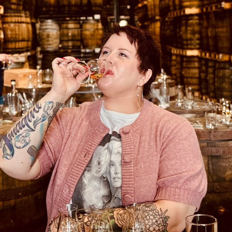 A owman with short hair and tattoos sipping from a class of whiskey