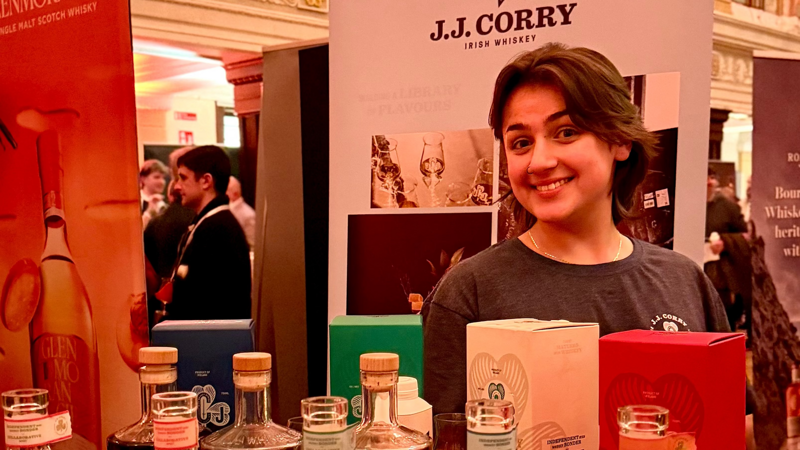 Woman standing behind a table with J.J. Corry whiskey bottles in an indoor setting