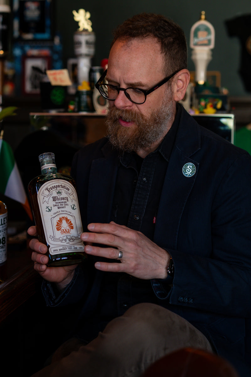 A man with a jacket and glasses holding a bottle of whiskey